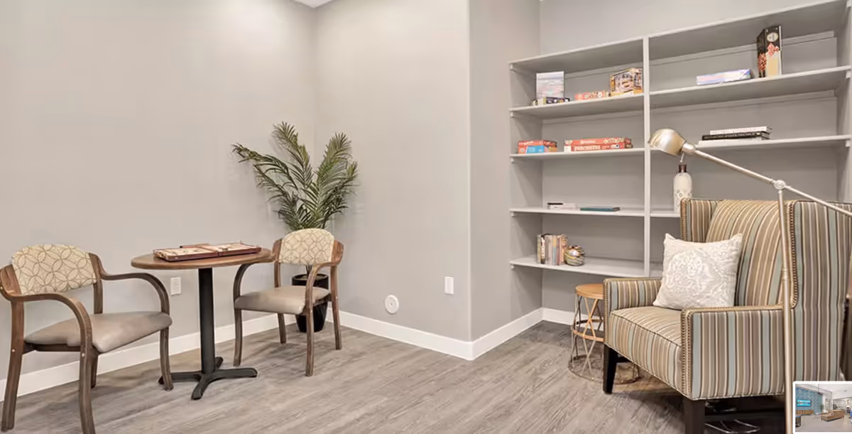 A cozy corner in a senior living facility featuring two cushioned chairs around a small round table with a board game on it, a potted plant in the corner, a striped armchair with a decorative pillow, a floor lamp, and a bookshelf with various board games and books.
