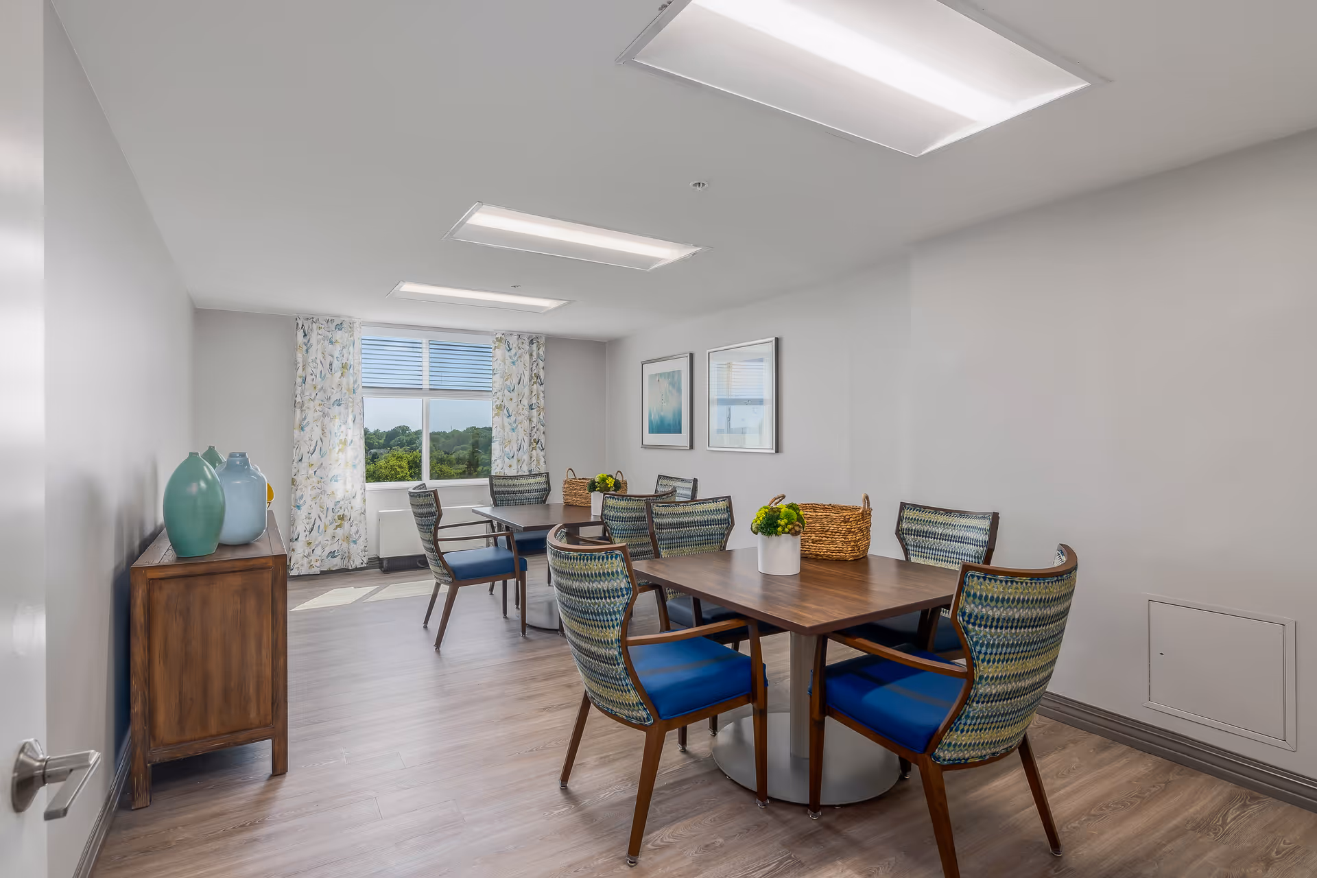 A bright and clean dining room with two wooden tables, each surrounded by four chairs with blue cushions and patterned backs. The room has light wood flooring, white walls, and a window with floral curtains letting in natural light. There are two framed artworks on the wall and a wooden sideboard with decorative vases on the left side.