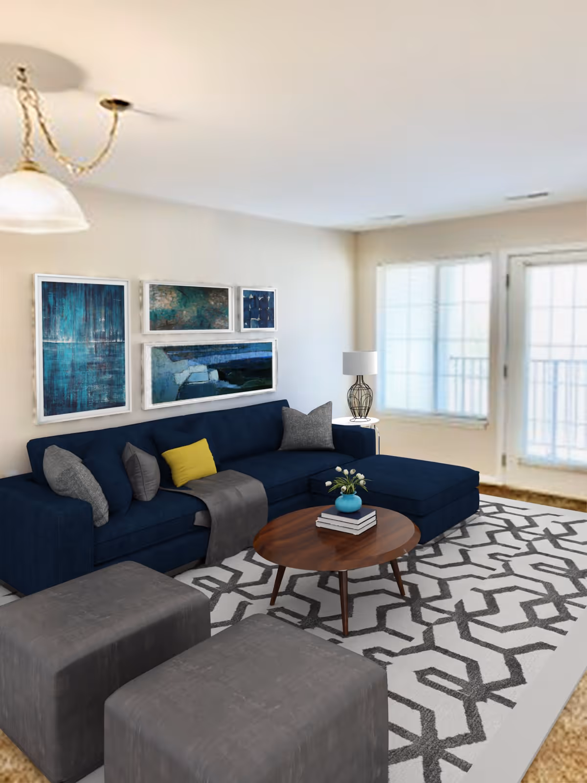 A modern living room with a dark blue sectional sofa adorned with gray and yellow pillows, a wooden coffee table with books and a small vase of flowers, two gray ottomans, a patterned area rug, a side table with a decorative lamp, and four abstract blue-themed paintings on the wall. Large windows and a glass door allow natural light into the room.