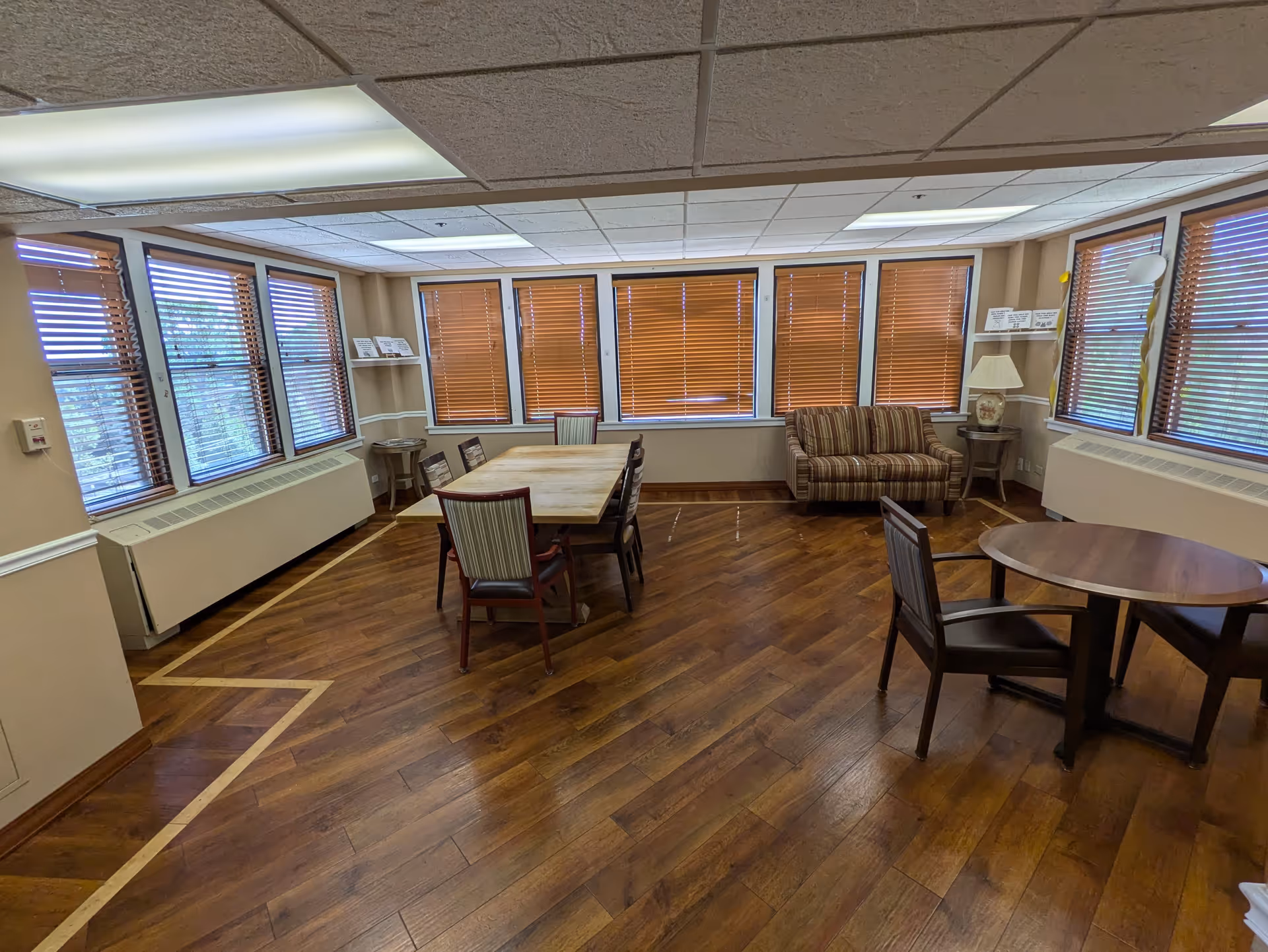 A well-lit room with wooden flooring and multiple windows with brown blinds. The room contains a rectangular wooden table with six chairs around it, a small round table with two chairs, a striped loveseat, a lamp on a side table, and some shelves with papers or books. The ceiling has fluorescent lighting panels.