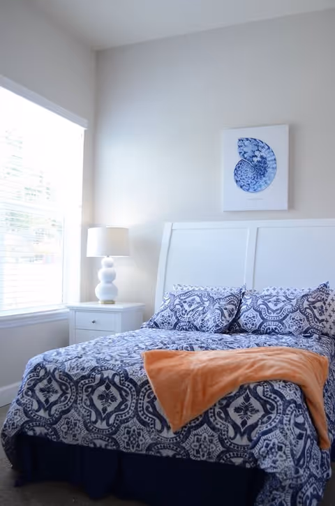 A bright bedroom with a large window on the left letting in natural light. The bed has a white headboard and is made with blue and white patterned bedding, with an orange throw blanket draped across the foot. A white nightstand beside the bed holds a white lamp with a round base. Above the bed hangs a framed artwork featuring a blue and white seashell design.