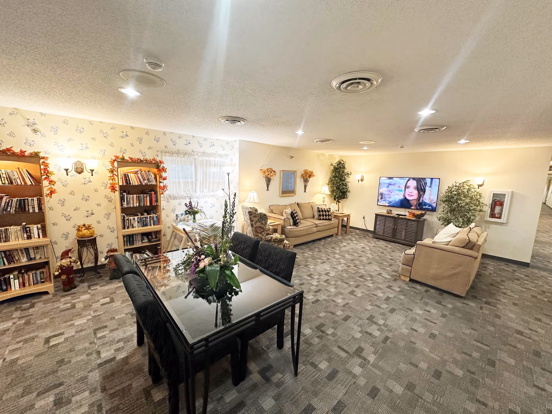A spacious living room area in an assisted living facility featuring two beige sofas, an armchair, a flat-screen TV mounted on a cabinet, and a glass dining table with black chairs. The room has carpeted flooring, floral wallpaper on one wall, bookshelves filled with books, decorative plants, and warm lighting from lamps and wall sconces.