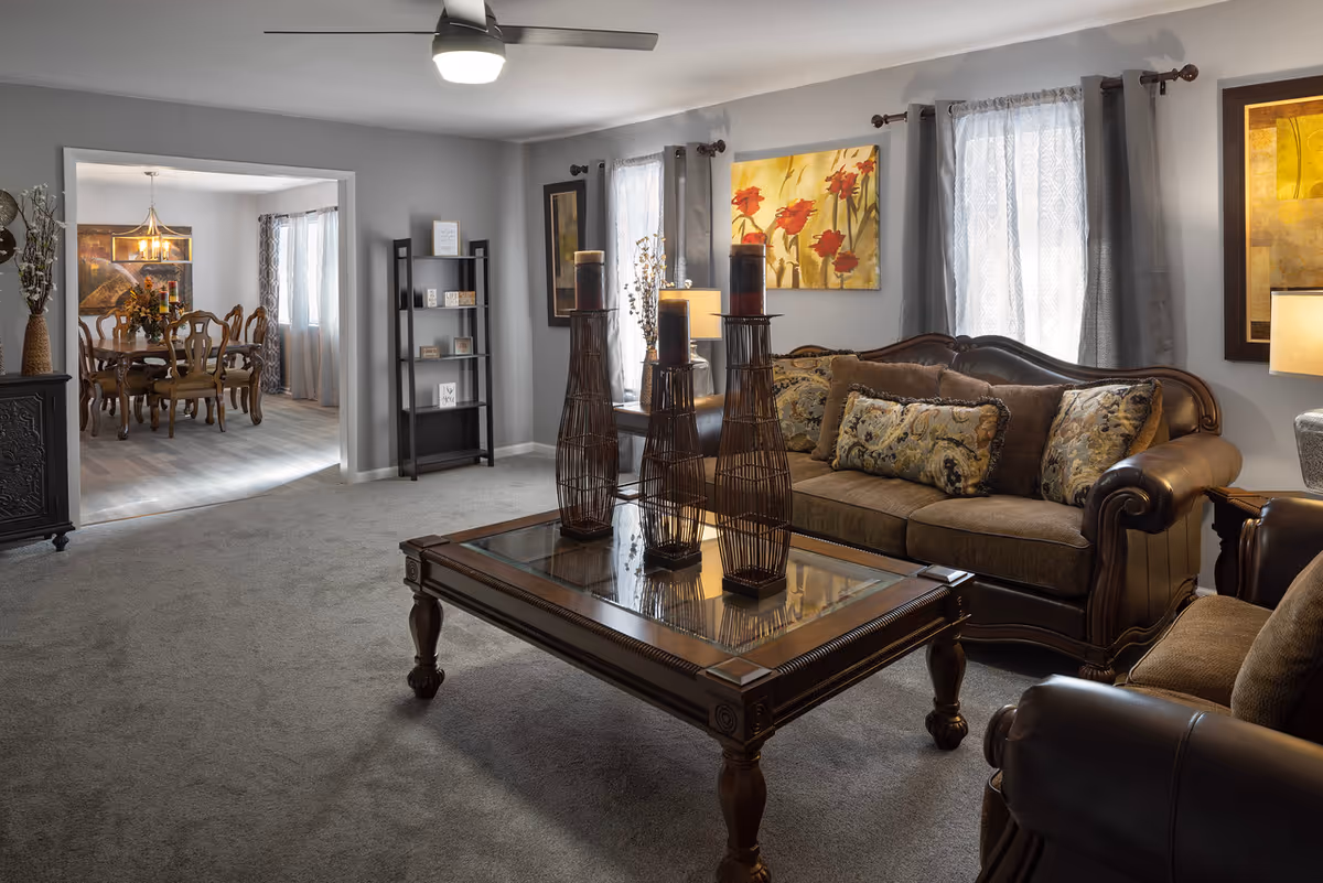 A cozy living room with upholstered sofas, patterned throw pillows, a glass-topped wooden coffee table with tall decorative candle holders, and a view into an adjoining dining room.