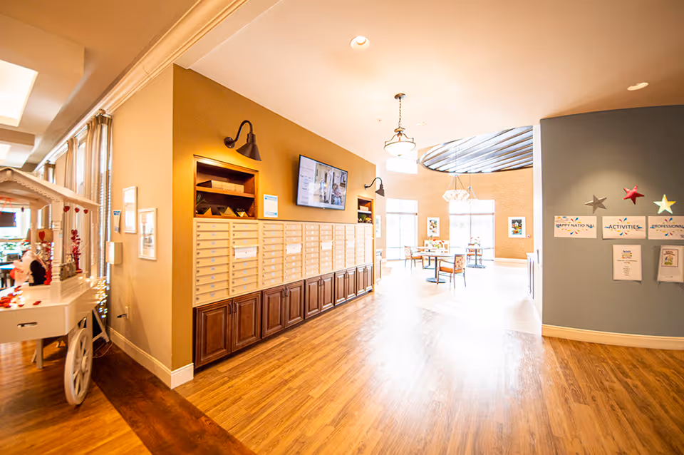 Interior view of a senior living facility hallway with a row of mailboxes mounted on a tan wall, a flat-screen TV above them, and a bright common area with tables and chairs in the background. The floor is wooden, and there are decorative stars and bulletin boards on a blue wall to the right.