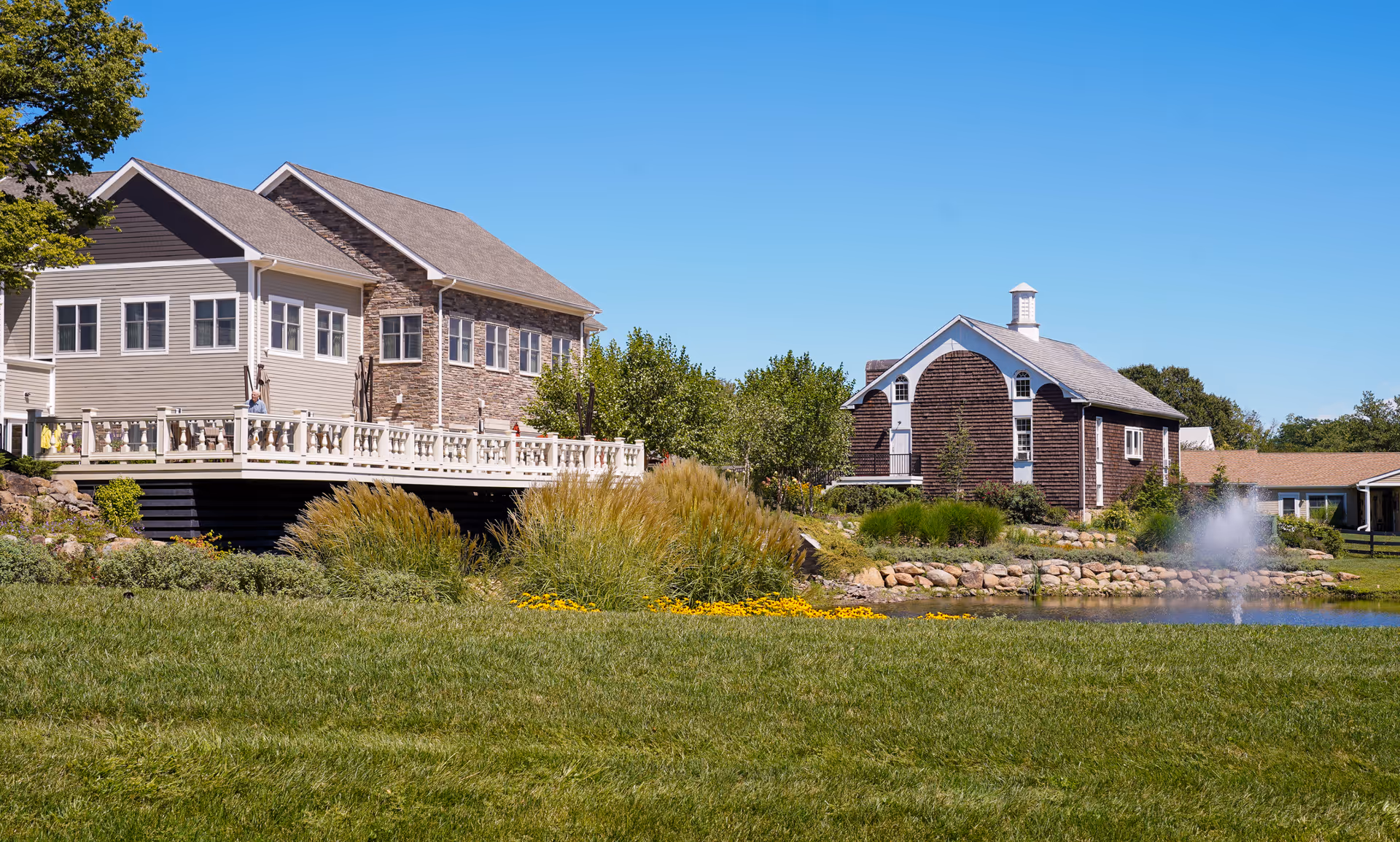 Exterior view of Pine Run Village showing a large building with beige siding and stone accents next to a smaller brown wooden building. There is a well-maintained grassy area, a pond with a water fountain, and landscaping with bushes and yellow flowers under a clear blue sky.
