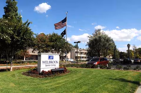 Exterior view of a senior living campus with a sign, flagpole, trees, and parked cars.