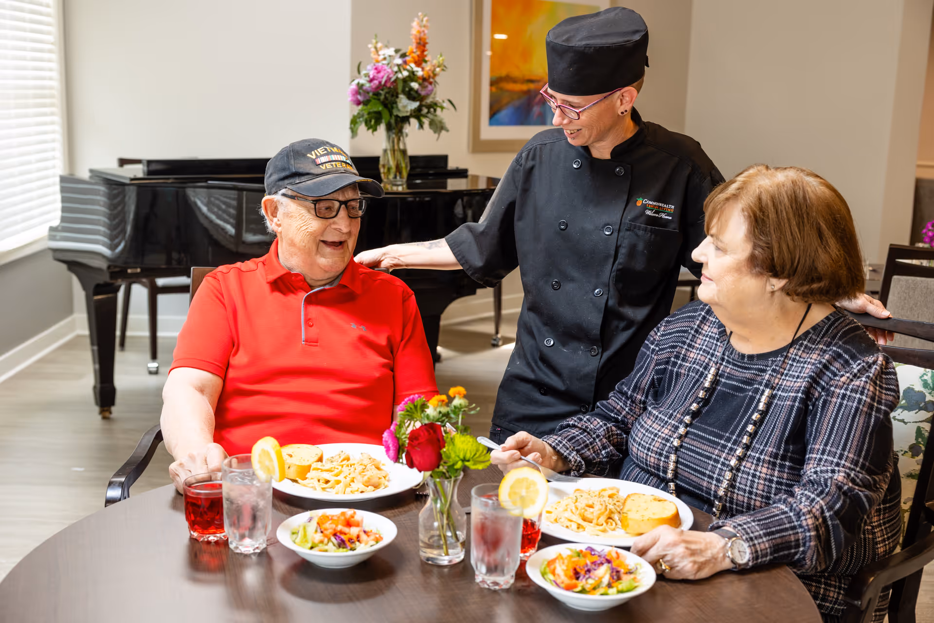 Two seniors seated at a dining table enjoying meals while a staff member in a chef uniform smiles and talks with them, with a piano in the background.