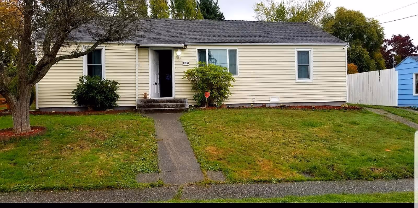 Single-story beige house with a dark shingled roof, a small front porch with steps, a concrete walkway leading to the front door, green lawn, a tree on the left side, and bushes near the entrance. A blue shed and white fence are visible on the right side.
