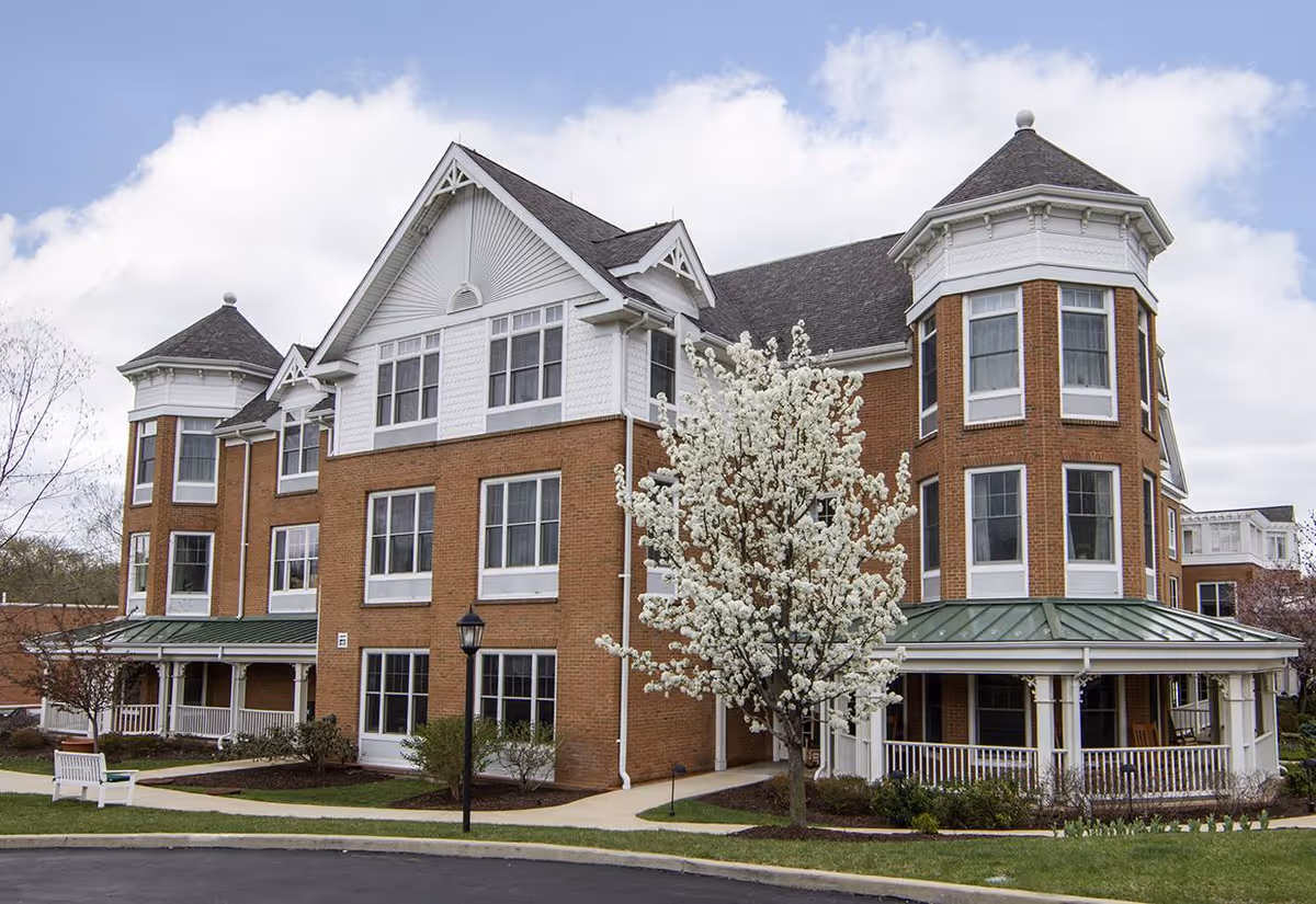Exterior view of a large, multi-story senior living facility building with brick and white siding. The building features multiple windows, two turret-like structures, a covered porch with white railings, and a blossoming tree in front. The sky is partly cloudy.