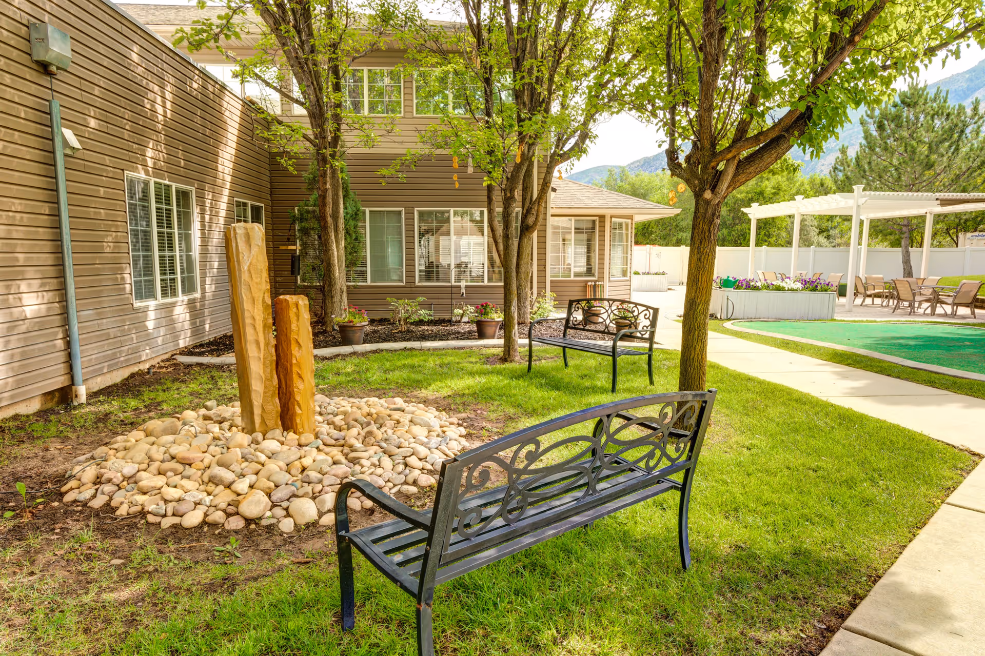 Outdoor garden area at The Auberge at North Ogden featuring two black metal benches, a small rock garden with two vertical stone sculptures, several trees, a paved walkway, and a covered seating area with chairs and tables in the background.