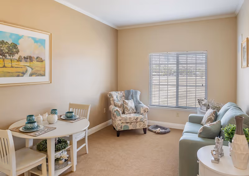 A cozy living room with beige walls and carpet, featuring a small round dining table set for two with teal dishes, a patterned armchair with a blue throw blanket, a light blue sofa with pillows, a window with blinds, and decorative items including plants and artwork on the walls.