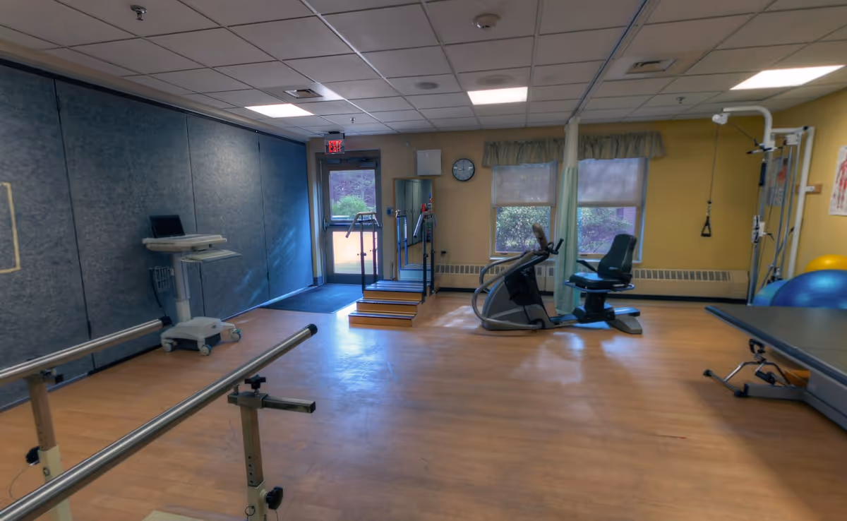 A rehabilitation or physical therapy room with wooden flooring, parallel bars for walking exercises, a small set of stairs with handrails, an exercise bike, therapy balls, and a treatment table. The room has yellow walls, two windows with curtains, and a door leading outside.