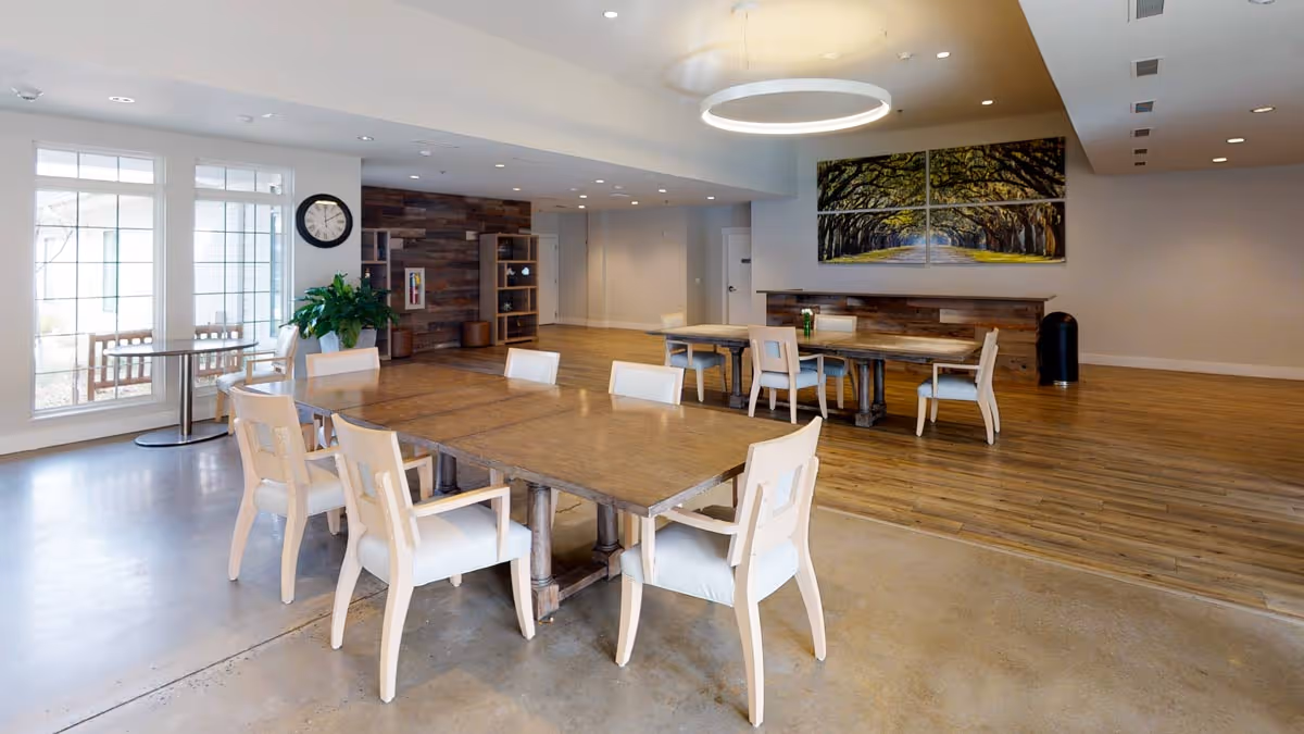 A spacious, well-lit common area with multiple wooden tables and white cushioned chairs arranged around them. The room features large windows on the left side, a modern circular ceiling light, a wall clock, a green plant, and a large multi-panel artwork depicting a tree-lined path on the far wall. The flooring transitions from polished concrete to wood.