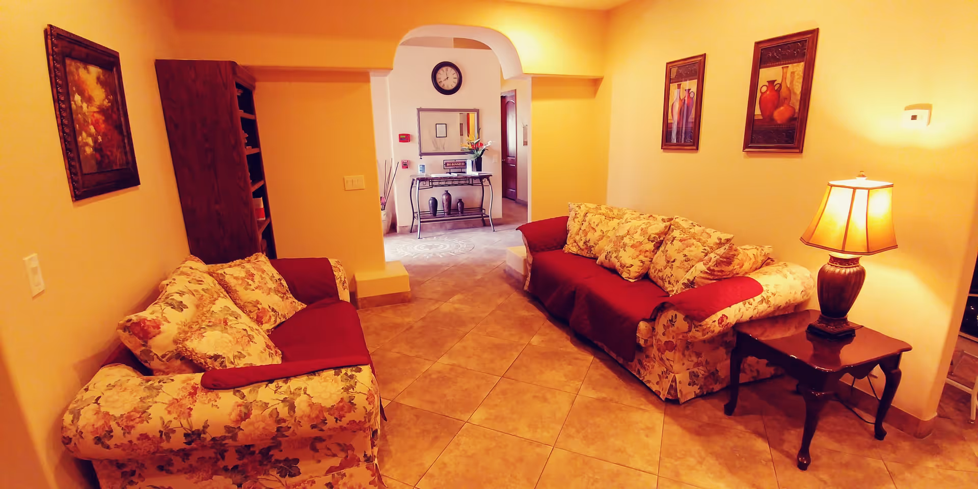 A cozy living room with two floral-patterned sofas covered partially with red throws, a wooden side table with a lamp, and framed artwork on the walls. The room has tiled flooring and an archway leading to another area with a clock and decorative items on a console table.