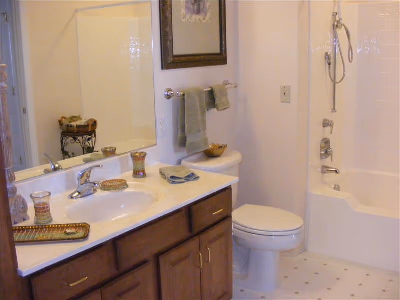 A clean bathroom with a white countertop sink, wooden cabinets, a toilet, and a bathtub with a showerhead. There are green towels hanging on a towel rack above the toilet and decorative items on the countertop and a small stand in the corner.