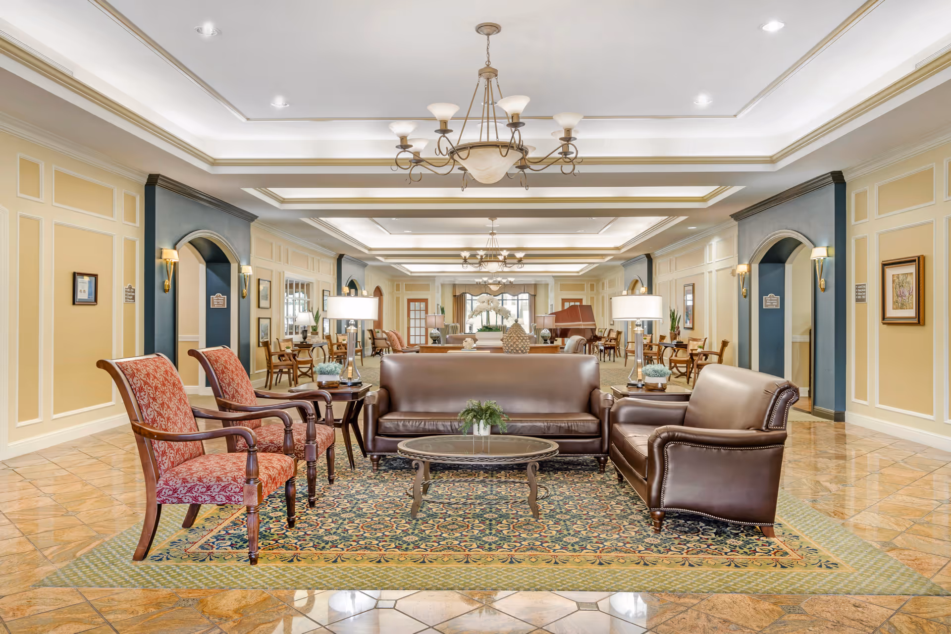 A spacious and elegant living room area with leather sofas, two upholstered armchairs, a glass coffee table on a patterned rug, and multiple chandeliers hanging from a coffered ceiling. The room features warm yellow walls with white trim, tiled floors, and several side tables with lamps and decorative plants. There are archways and framed artwork along the walls, creating a welcoming and comfortable atmosphere.