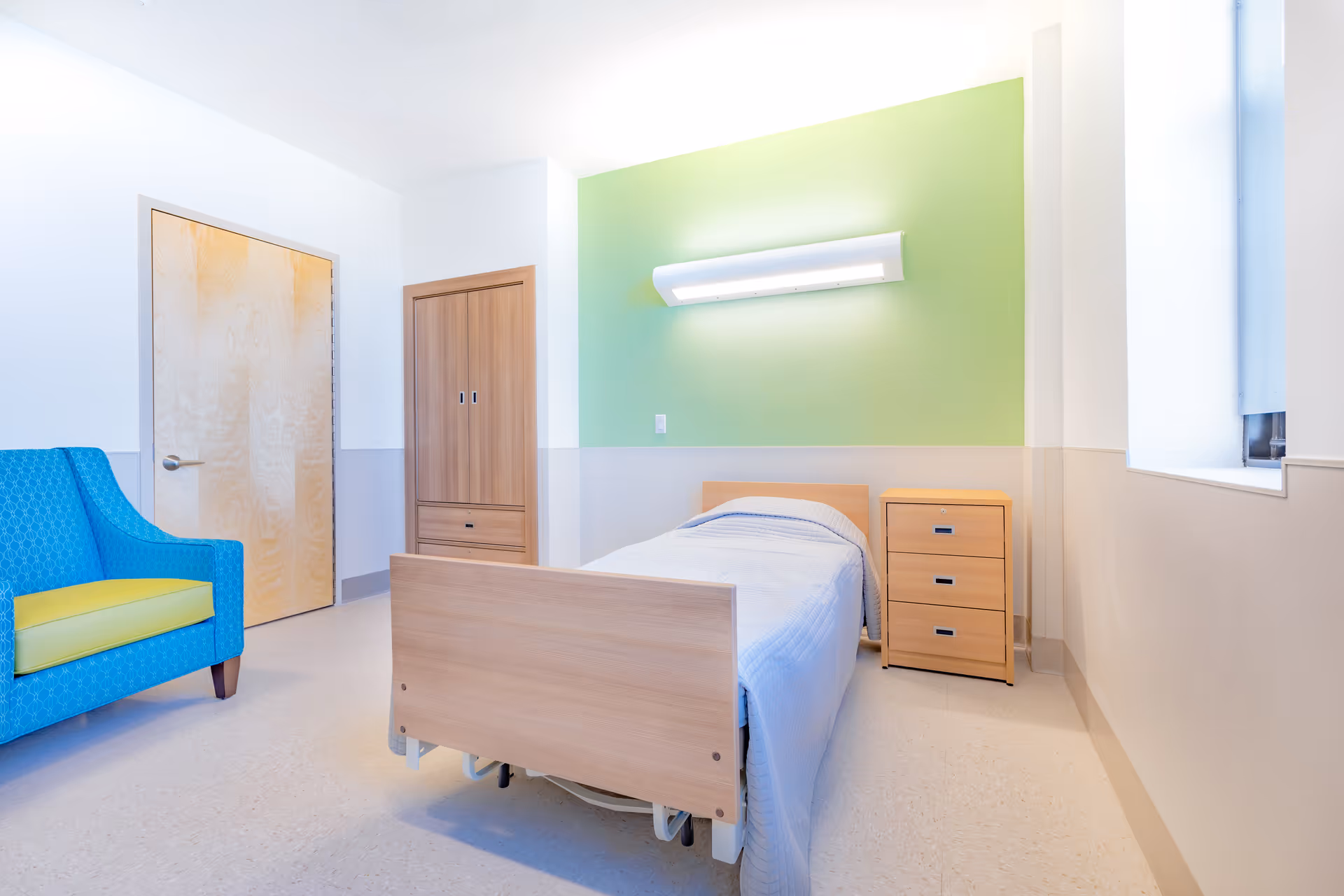 A simple private bedroom with a single hospital-style bed against a green accent wall, a wooden nightstand and wardrobe, and a blue armchair with a yellow cushion.