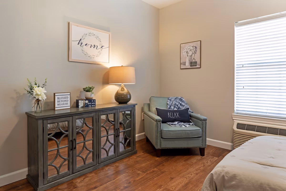 A cozy corner of a senior living bedroom featuring a green armchair with a 'RELAX.' pillow and a plaid throw blanket. Next to the chair is a wooden cabinet with decorative items including flowers, a lamp, and framed signs. The wall has two framed pictures, one with the word 'home'. A window with blinds and an air conditioning unit is visible on the right side.