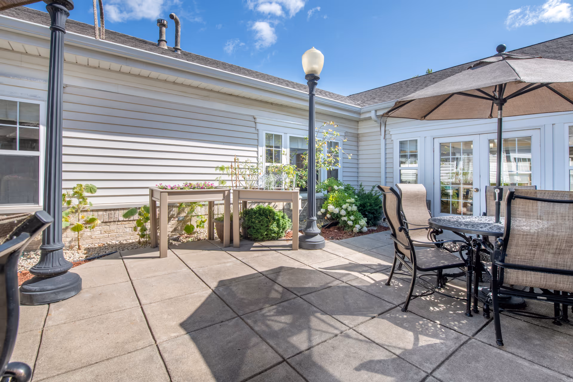 Outdoor patio area at Brookdale Plymouth with a round metal table and four chairs under a large umbrella. The patio is paved with square tiles and has two black lamp posts. There are plants and flowers along the building wall, and the sky is clear and blue.