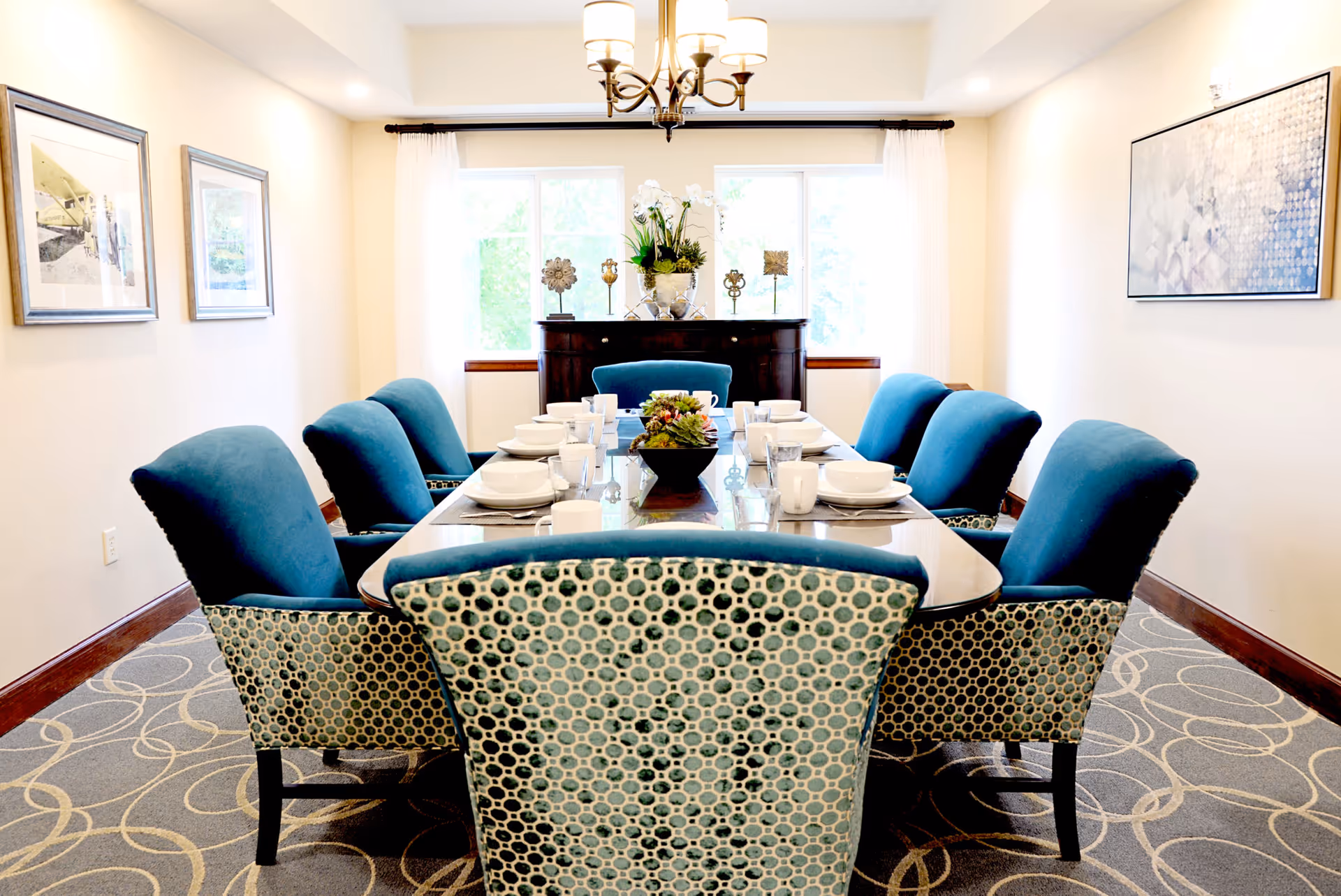 A bright dining room with a long glass-top table set for eight people with white dishes, cups, and glasses. The chairs have blue cushions and patterned backs. There is a chandelier overhead, two framed pictures on the left wall, a large abstract painting on the right wall, and a sideboard with decorative items and a flower arrangement in front of two large windows with white curtains.