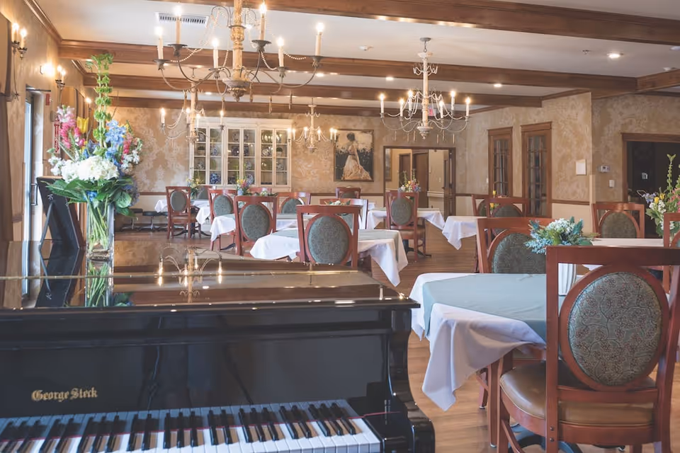 Elegant dining room with a grand piano in the foreground, multiple tables with white tablecloths, chandeliers, floral arrangements, and wooden chairs.
