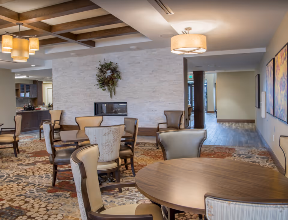 A furnished communal dining/lounge area with round wooden tables, upholstered chairs, a stone fireplace wall and pendant lighting.