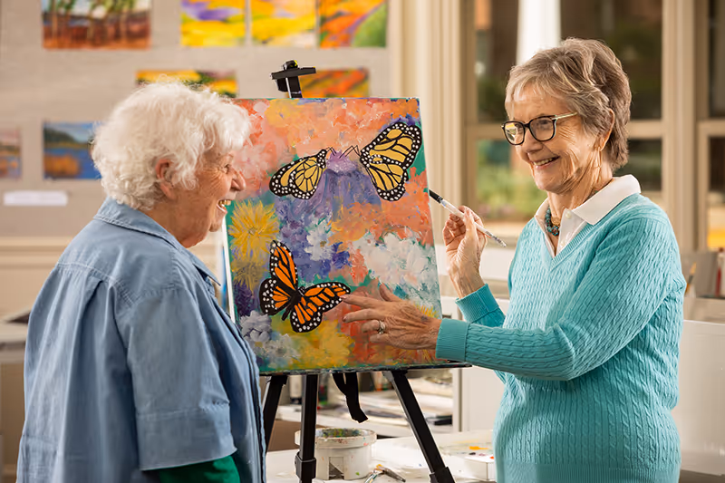 Two elderly women smiling and engaging in painting a colorful canvas featuring butterflies and flowers in a well-lit art room with paintings displayed on the walls.