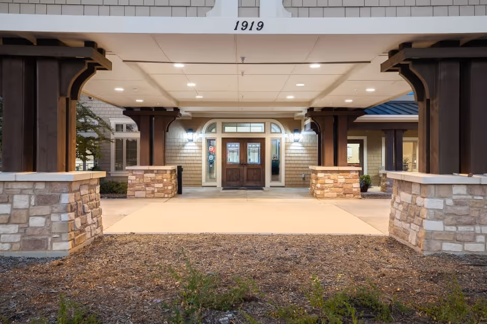 Front entrance and covered porte-cochere of a senior living building with stone pillars and double wooden doors under the address number 1919.
