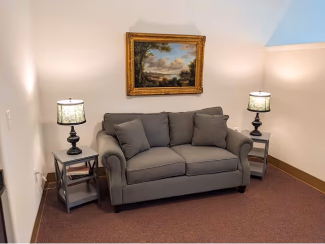 A cozy sitting area with a gray loveseat sofa flanked by two matching side tables, each with a decorative lamp. Above the sofa hangs a framed landscape painting with a gold ornate frame. The walls are light-colored and the floor is carpeted in a muted tone.