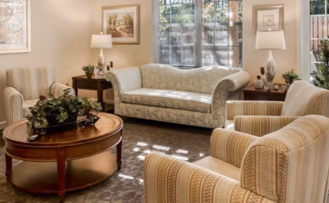 A cozy living room with a patterned sofa and two striped armchairs arranged around a wooden coffee table with a plant centerpiece. The room has large windows with blinds letting in natural light, two side tables with lamps, and framed artwork on the walls.