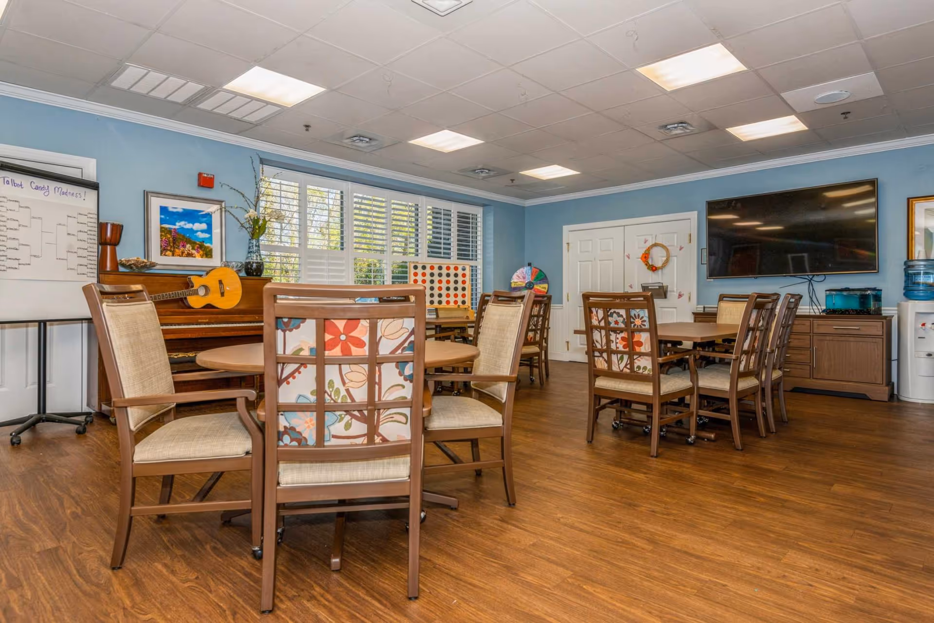 A well-lit activity room with wooden floors and light blue walls. The room contains several wooden tables surrounded by chairs with floral and beige upholstery. A piano with a guitar resting on it is positioned against one wall, next to a whiteboard labeled 'Talbot Candy Madness!'. There is a large flat-screen TV mounted on the wall, a water cooler, and a colorful game wheel in the corner. Large windows with white shutters allow natural light to enter the room.