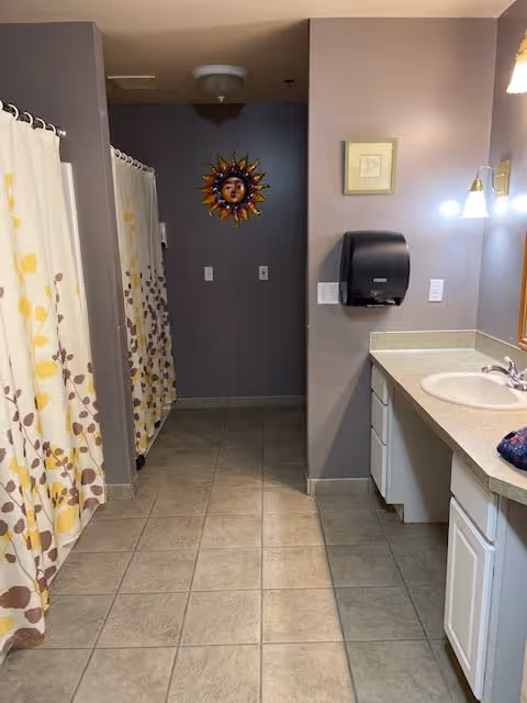 Interior view of a bathroom area with two shower stalls on the left, each with a curtain featuring a leaf pattern. On the right side, there is a countertop with a sink, a wall-mounted soap dispenser, and a mirror with lights above it. The walls are painted gray, and there is a decorative sun face hanging on the back wall.
