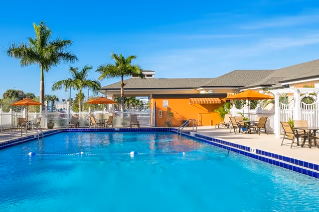 Outdoor swimming pool area with clear blue water, surrounded by palm trees, patio tables with orange umbrellas, and a building with a gray roof and orange wall under a clear blue sky.