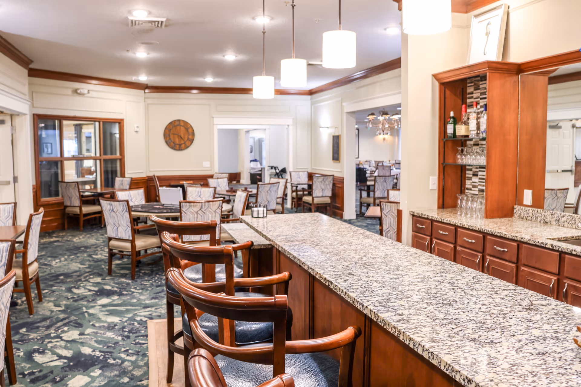 Interior view of a senior living facility dining area with multiple tables and chairs arranged neatly. In the foreground, there is a granite countertop bar with wooden bar stools. The room features warm lighting with pendant lights hanging from the ceiling, wood paneling, and a decorative wall clock.