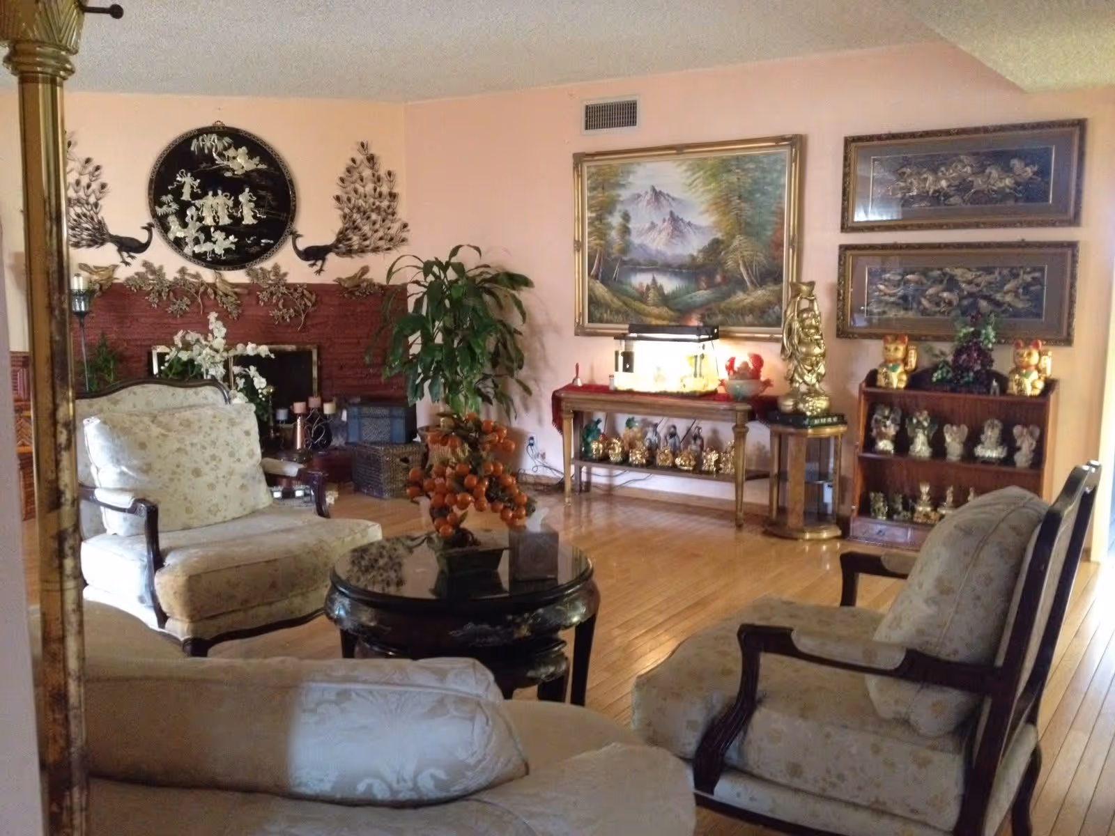 A living room with floral upholstered armchairs and a round black coffee table with an orange fruit centerpiece. The room features a red brick fireplace decorated with metal peacock wall art and a round black and gold decorative plate. There is a large green plant next to the fireplace. On the opposite wall, there is a large framed painting of a mountain landscape and two smaller framed artworks above a wooden table and bookshelf displaying various gold and ceramic figurines, including lucky cat statues. The floor is wooden, and the walls are painted light pink.