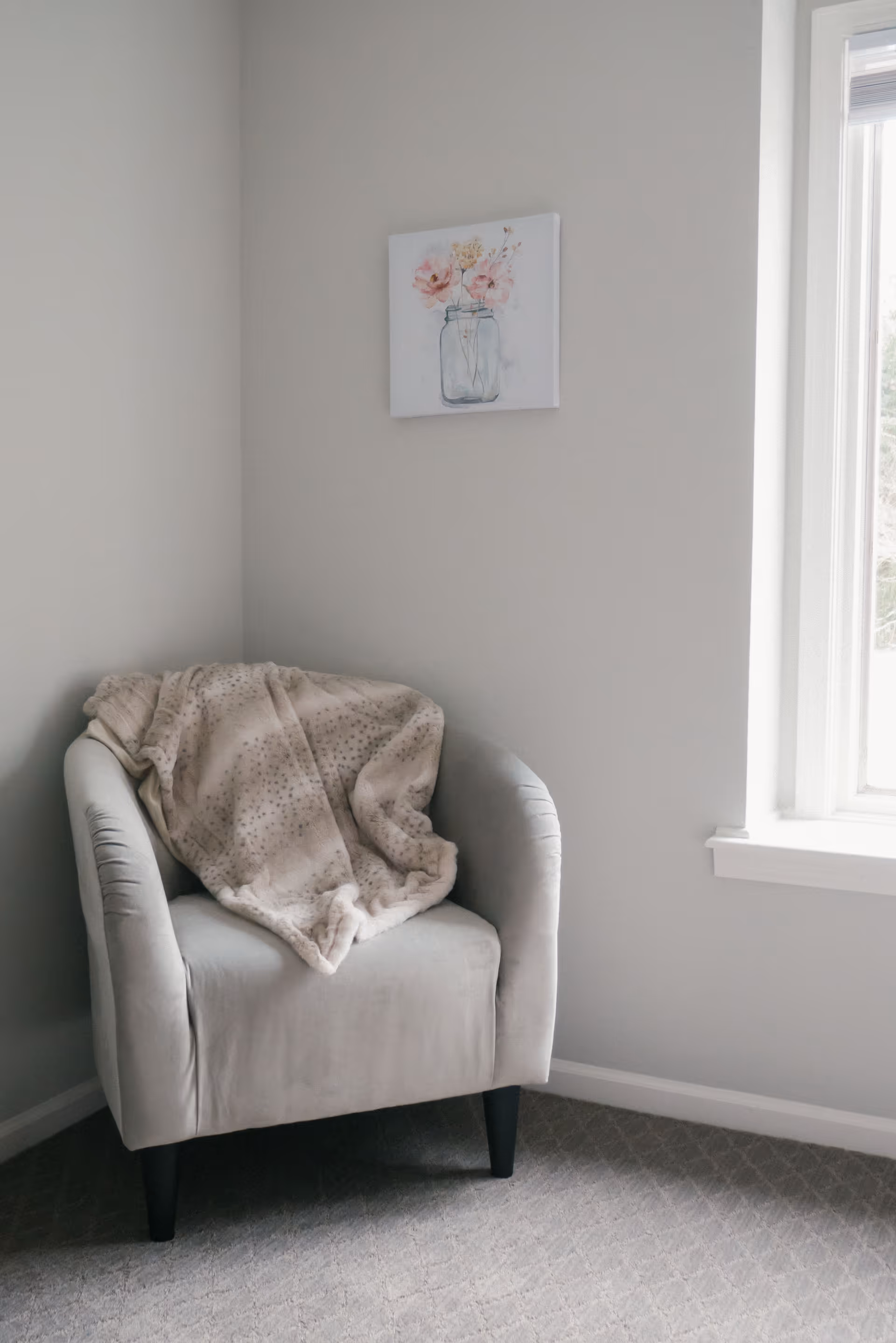 A cozy corner with a light gray upholstered armchair draped with a soft, beige spotted blanket. Above the chair hangs a small painting of pink flowers in a glass jar. To the right, there is a window letting in natural light, and the walls and carpet are in neutral tones.