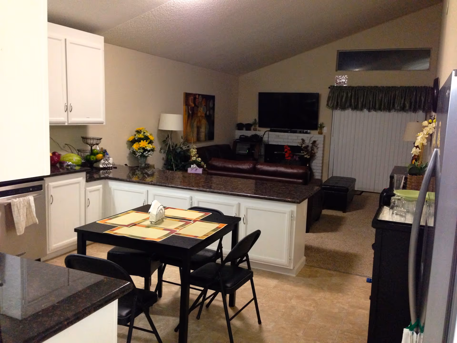 Interior view of a residential care facility showing a kitchen area with white cabinets, a black dining table with four chairs and placemats, and a living room in the background with a brown leather sofa, a flat-screen TV mounted on the wall, a fireplace, and a sliding glass door with vertical blinds and a green valance.