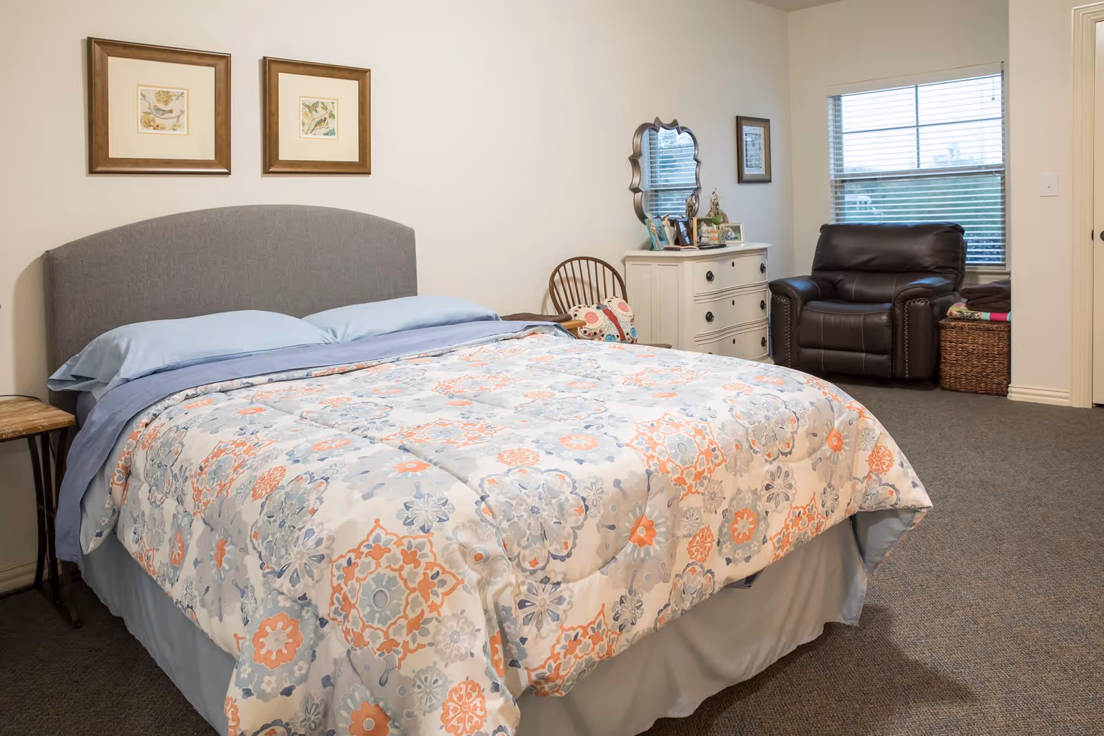 A bedroom with a large bed covered in a floral patterned comforter in shades of blue, orange, and white. The bed has a gray upholstered headboard and blue pillows. On the left side of the bed is a small wooden side table. Against the far wall is a white dresser with a decorative mirror and framed photos on top. Next to the dresser is a wooden chair with a colorful cushion. In the corner near a window with blinds is a dark brown leather armchair and a woven basket. The room has beige walls and carpeted flooring.