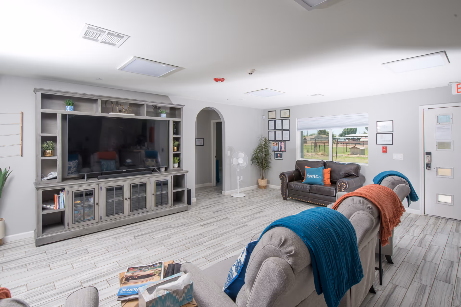 A bright and modern living room in an assisted living facility featuring a large flat-screen TV on a wooden entertainment center, two gray armchairs with blue and orange throws, a brown leather loveseat with decorative pillows, a standing fan, and a window showing an outdoor view.