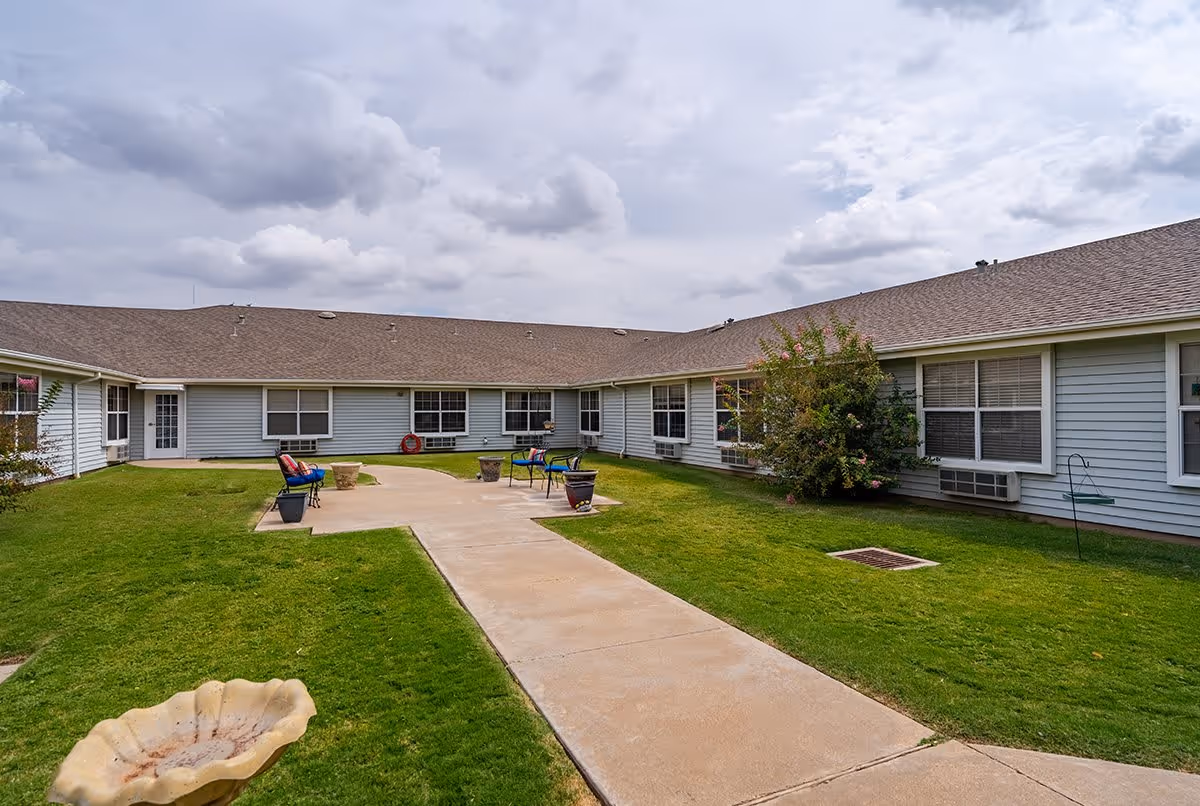 Outdoor courtyard area of Mackenzie Court facility with a concrete walkway, green grass, several chairs with cushions, potted plants, and a cloudy sky overhead. The building surrounds the courtyard with multiple windows and a door visible.