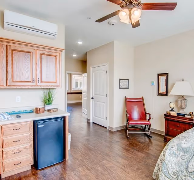 Furnished bedroom interior with wooden cabinets and mini-fridge, a red rocking chair, nightstand with lamp, and a ceiling fan.