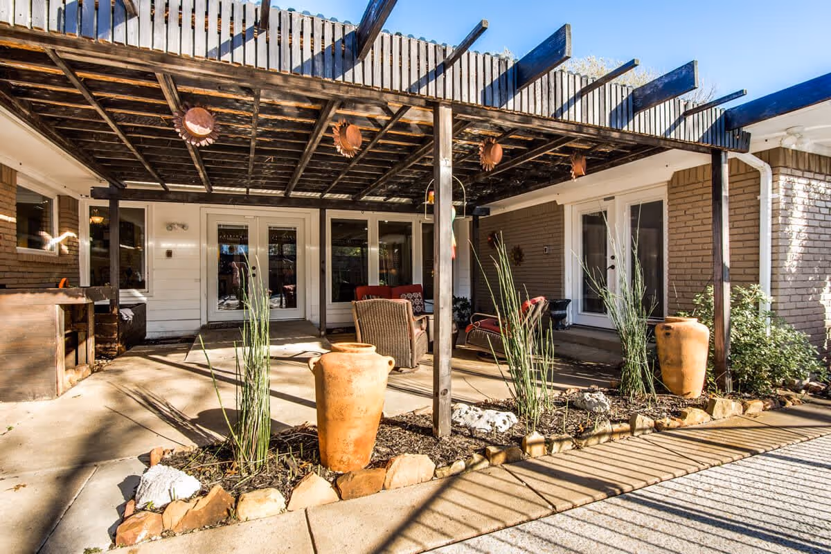 Outdoor patio area at Sage Oak - Lake Highland with a wooden pergola overhead, two large terracotta pots with tall plants, wicker chairs with cushions, and glass doors leading inside the building.