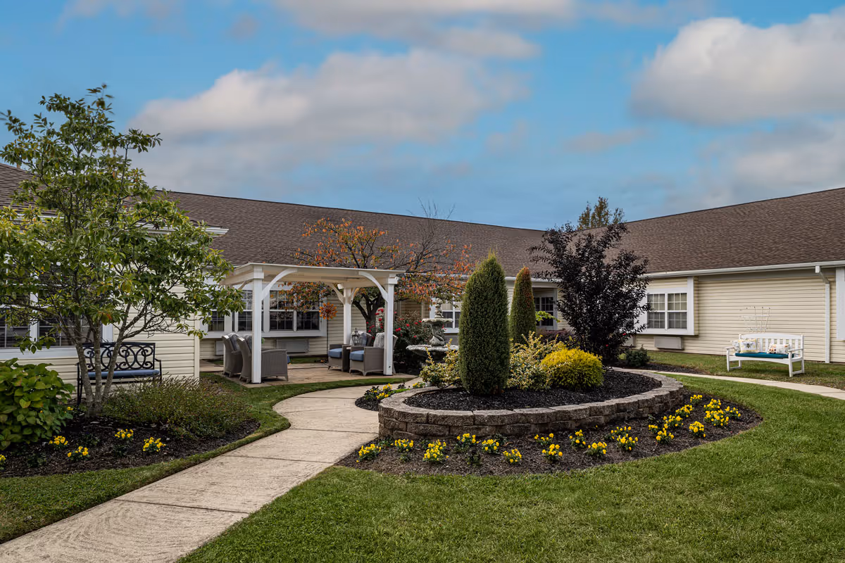 Outdoor courtyard area of a senior living facility with a paved walkway, landscaped garden beds with yellow flowers and shrubs, a white pergola with seating underneath, and benches placed around the grassy area. The building surrounds the courtyard with beige siding and multiple windows under a brown roof, under a partly cloudy blue sky.