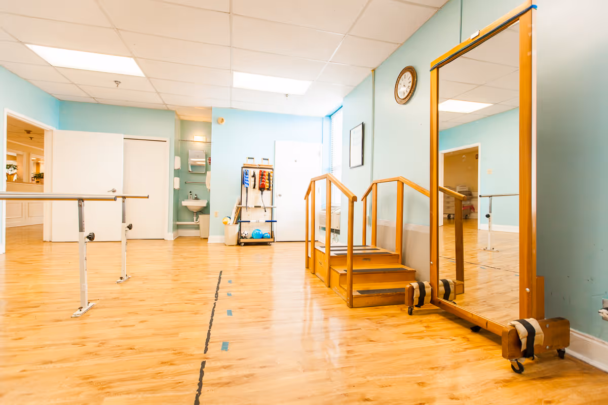 A spacious room with light blue walls and wooden flooring featuring physical therapy equipment including parallel bars, a small wooden staircase with handrails, a large mirror on wheels, and a rack holding exercise balls and resistance bands. There is a clock on the wall and a small sink in the corner.