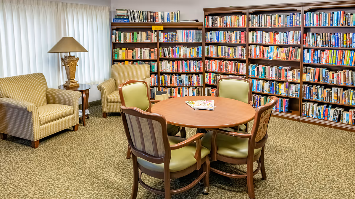 Cozy reading room with a round table and chairs, armchairs, lamp, and bookshelves lining the wall.