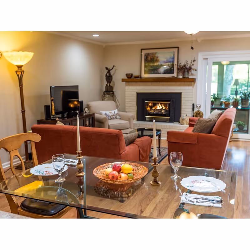Cozy living room and dining area with a glass dining table set in the foreground, red sofas, and a lit fireplace.