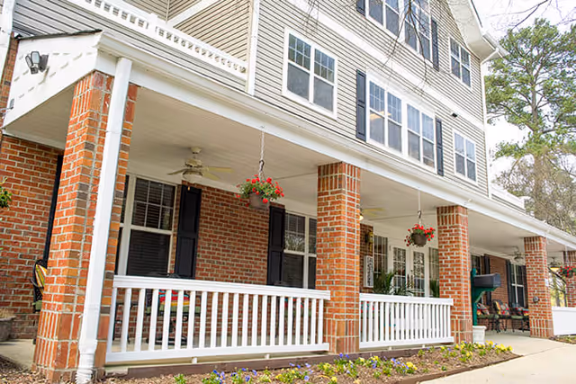 Exterior view of a senior living facility with a covered porch supported by brick columns and white railings. Hanging flower baskets and ceiling fans are visible on the porch, with windows and a landscaped flower bed in front.