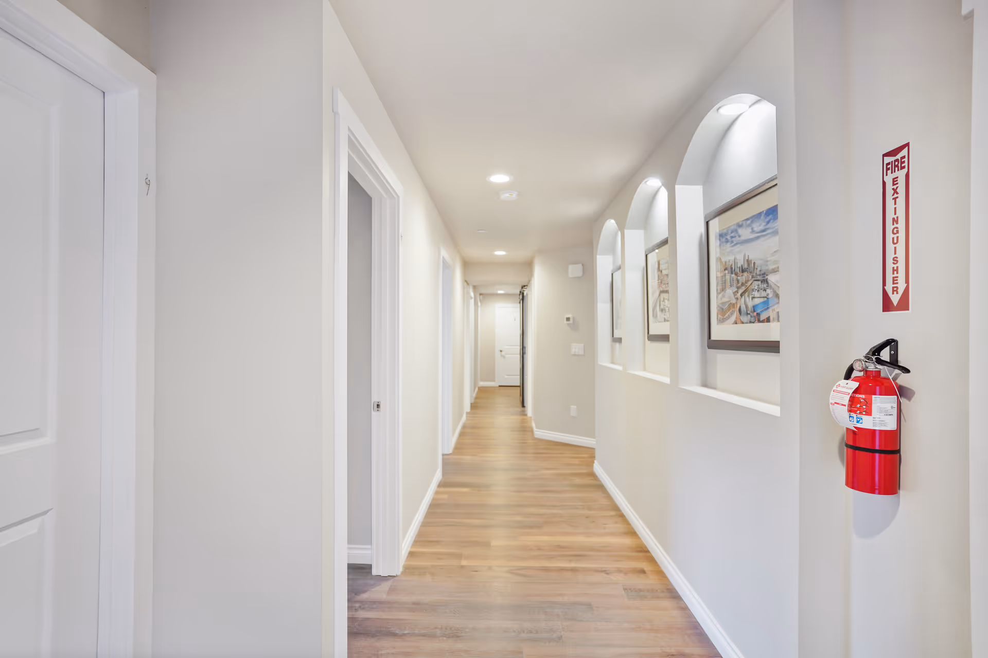 Long, well-lit interior hallway with wood flooring, doors on the left, arched display openings and framed art on the right, and a fire extinguisher mounted on the wall.