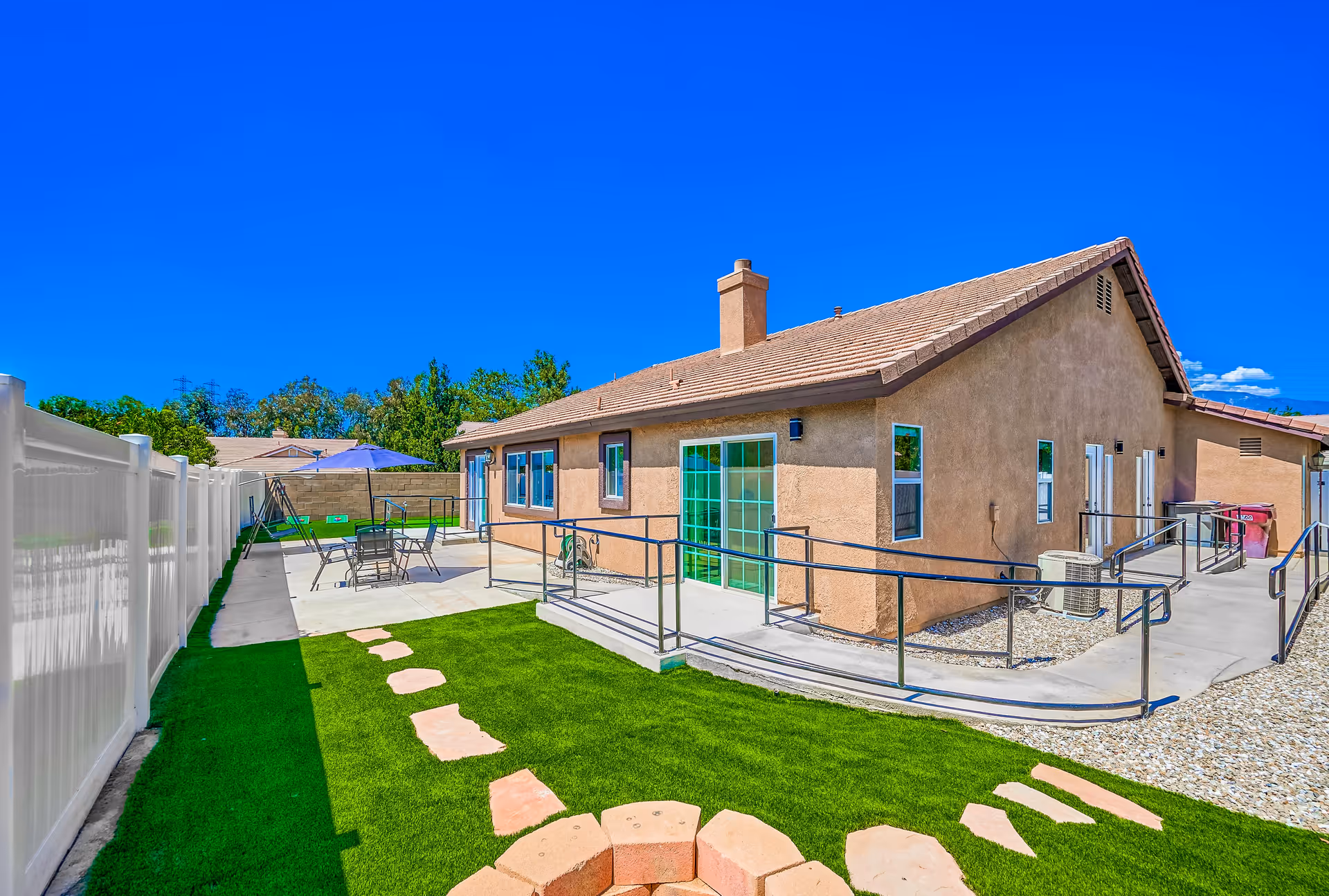 Outdoor view of a single-story building with a tan stucco exterior and a tiled roof under a clear blue sky. The area features a concrete patio with a table, chairs, and an umbrella, a green lawn with stepping stones, a fire pit, and metal handrails along the walkways. A white fence runs along one side of the property.