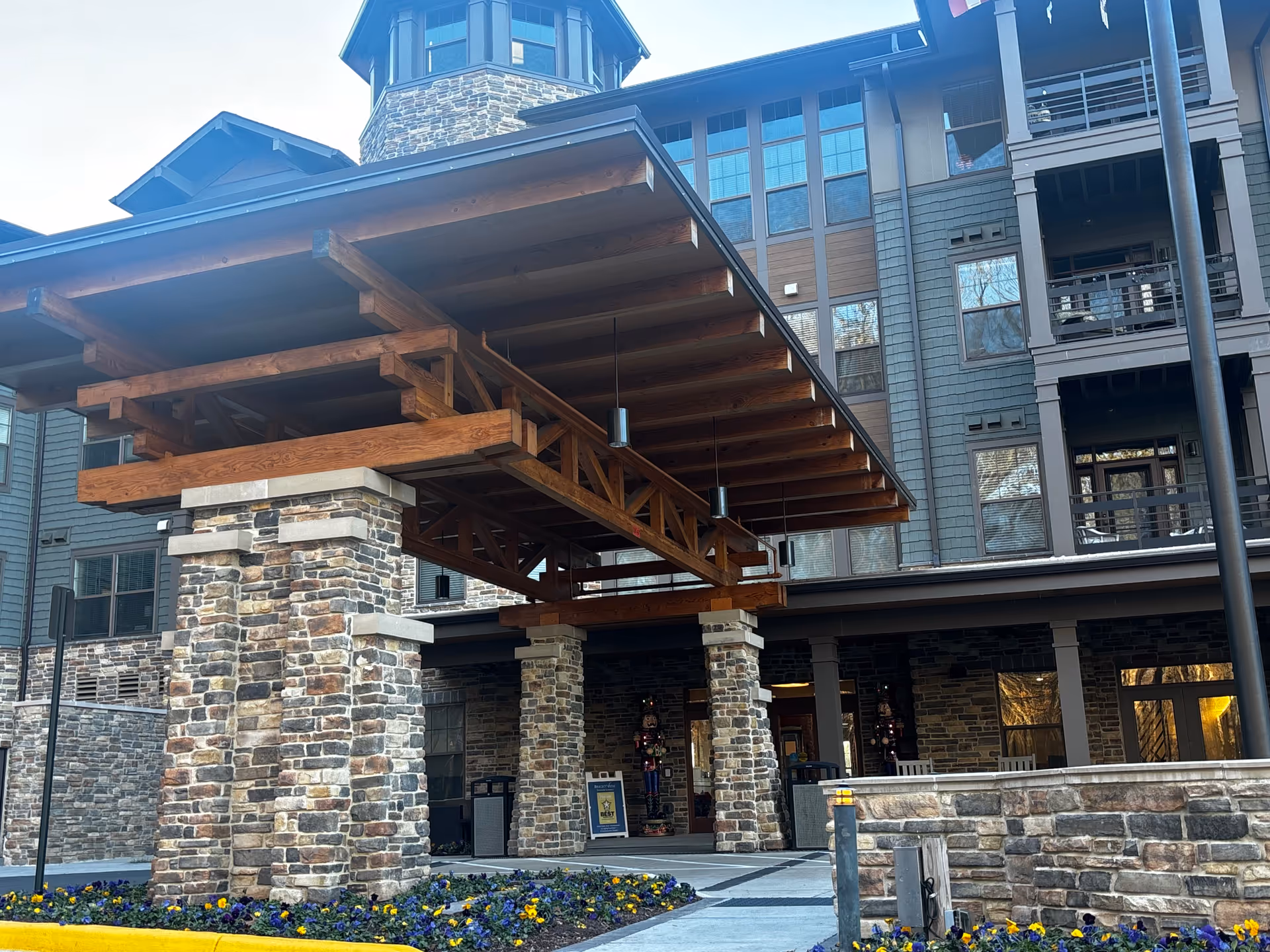 Entrance of a senior living facility with a large wooden and stone covered driveway supported by stone pillars. The building exterior features multiple windows and balconies, with flower beds containing purple and yellow flowers in front.